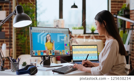 Asian graphic designer uses editing software for photo retouching in a well organized home workspace. Female freelancer uses digital tools essential for a creative artistic career. Camera B. 125638234