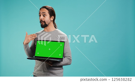 Tech content creator doing influencer marketing using green screen laptop, isolated over studio background. Arab man holding empty copy space mockup notebook, doing review, camera B Tech content creator doing influencer marketing using green screen laptop, isolated over studio background. Arab man holding empty copy space mockup notebook, doing review, camera B 125638282