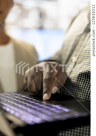 African american man next to colleagues in office coordinating company administrative operation, writing documents on notebook. Close up of employee typing on laptop keyboard, inputting data metrics 125638312