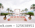 Long festive table with bouquets of flowers stands near a wicker sculpture in front of a pool with green palm trees on the sides 125638951