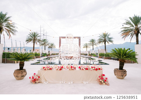 Long festive table with bouquets of flowers stands near a wicker sculpture in front of a pool with green palm trees on the sides Long festive table with bouquets of flowers stands near a wicker sculpture in front of a pool with green palm trees on the sides 125638951