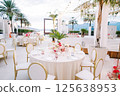 Festive tables with bouquets of flowers and white chairs stand near a stage decorated with garlands of light bulbs 125638953