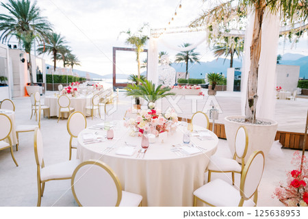 Festive tables with bouquets of flowers and white chairs stand near a stage decorated with garlands of light bulbs 125638953