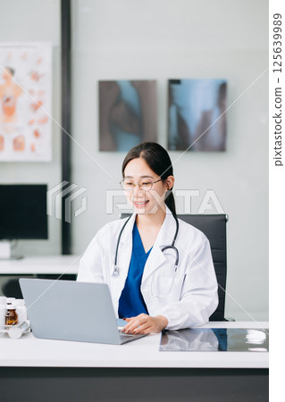 Confident young Asian female doctor in white medical uniform sit at desk working on computer. Smiling use laptop write in medical journal 125639989