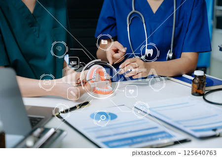Concentrated dentist sitting at table with jaw samples tooth model and working with tablet and laptop in dental office Concentrated dentist sitting at table with jaw samples tooth model and working with tablet and laptop in dental office 125640763