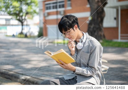 Asian man with glasses is smiling while reading a book with yellow cover, sitting on sidewalk bench. Asian man with glasses is smiling while reading a book with yellow cover, sitting on sidewalk bench. 125641488