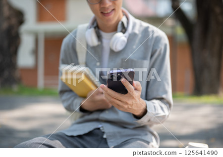 Close up of an asian man with glasses smiling while looking at his phone, sitting on sidewalk bench. 125641490