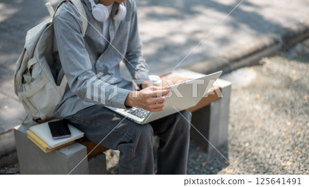 Close up of an asian man touching or adjusting his laptop screen, sitting on the sidewalk bench. 125641491