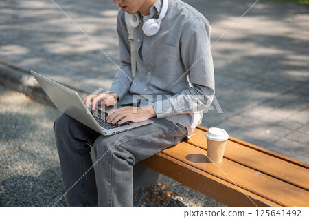 Close up of an asian man typing on his laptop, sitting on sidewalk bench. Study or working outdoor. 125641492