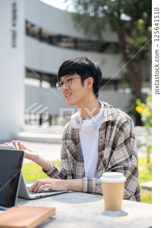 A smiling asian man with glasses explaining to a friend while sitting with laptop at table in park. 125641510