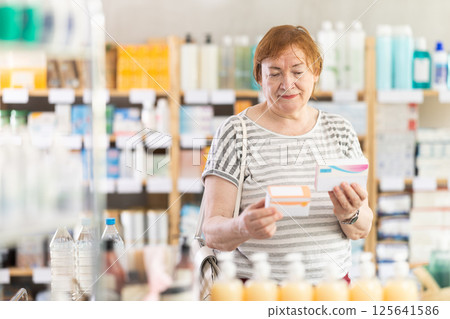 In sales area of pharmacy, mature woman examines packaging of tablet pill In sales area of pharmacy, mature woman examines packaging of tablet pill 125641586