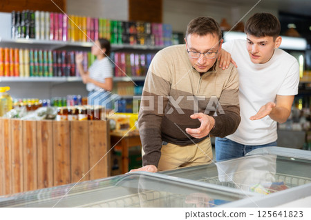 Man with glasses and young guy together chooses frozen food in fridge in supermarket 125641823