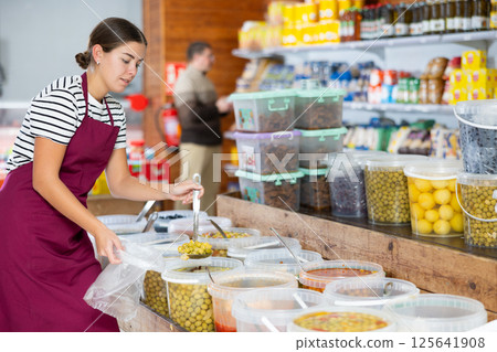 Young groceress scooping marinated olives into bag for customer at store Young groceress scooping marinated olives into bag for customer at store 125641908