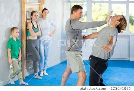 Trainer and man in group self-defense classes practicing sparring technique of blowing to chin in gym, in background family watches of performing technique 125641955