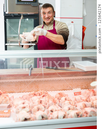 Meat department worker stands behind counter and holds chicken carcass in his hands Meat department worker stands behind counter and holds chicken carcass in his hands 125642174