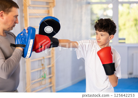 Teenage boy training boxing kicks on punch mitts held by instructor 125642253