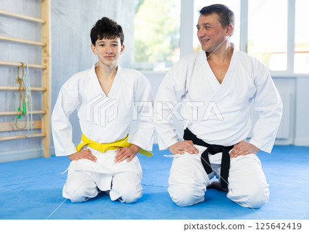 Teenage boy and middle-aged man wearing kimono sitting in seiza position in sports hall Teenage boy and middle-aged man wearing kimono sitting in seiza position in sports hall 125642419