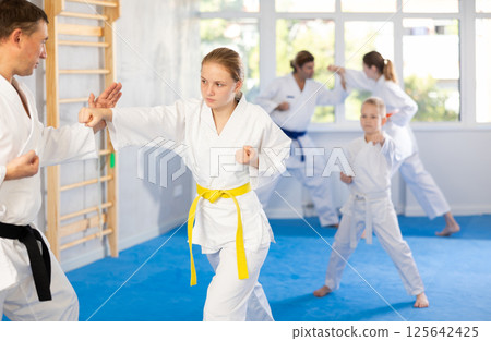 Young girl in kimono and colored belt practicing karate punch block during group martial arts lesson in gym, accompanied by trainer Young girl in kimono and colored belt practicing karate punch block during group martial arts lesson in gym, accompanied by trainer 125642425
