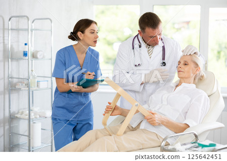 Man dermatologist examines skin of senior womans face, female nurse records data in client card Man dermatologist examines skin of senior womans face, female nurse records data in client card 125642451