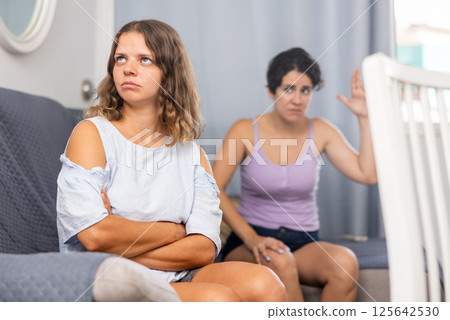 Upset young woman sitting at sofa with disgruntled woman behind Upset young woman sitting at sofa with disgruntled woman behind 125642530
