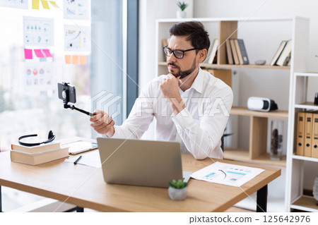 Professional businessman mid thirties with glasses seated at office desk filming video call or vlog with smartphone. Bright modern workspace with shelves in background 125642976