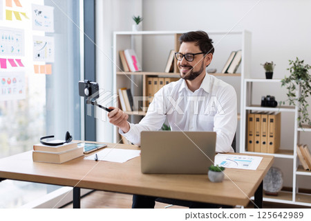 Young adult male recording video call, sitting by laptop in office environment, wearing glasses, business shirt, engaging with camera. Modern office setup with documents, tablet, and greenery Young adult male recording video call, sitting by laptop in office environment, wearing glasses, business shirt, engaging with camera. Modern office setup with documents, tablet, and greenery 125642989