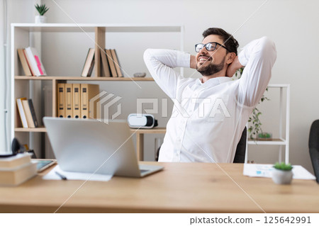 Young adult white male wearing glasses and white shirt sitting at desk smiling during office break, showcasing relaxation and positive work-life balance. 125642991