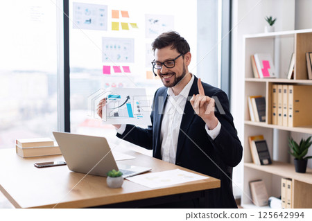 Young adult male business professional presenting charts and financial data in office. Confident and engaged person demonstrating growth metrics using visual aids, representing focus on progress Young adult male business professional presenting charts and financial data in office. Confident and engaged person demonstrating growth metrics using visual aids, representing focus on progress 125642994