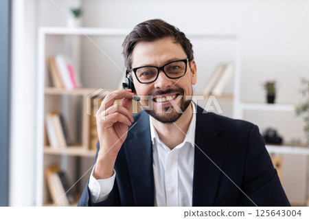 Confident adult Caucasian man wearing glasses and headset in modern office environment engaged in communication. 125643004