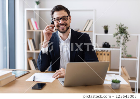 Confident adult Caucasian man wearing glasses and headset in modern office environment engaged in communication. Confident adult Caucasian man wearing glasses and headset in modern office environment engaged in communication. 125643005