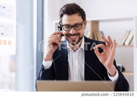 Businessman wearing headset conducting virtual meeting, gesturing okay sign, modern office background 125643010