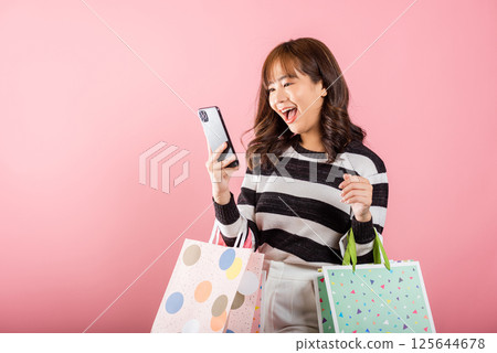 Portrait Asian happy beautiful young woman shopper smiling standing excited holding online shopping bags online multicolor and smartphone on hand, isolated on pink background, glad with mobile phone Portrait Asian happy beautiful young woman shopper smiling standing excited holding online shopping bags online multicolor and smartphone on hand, isolated on pink background, glad with mobile phone 125644678