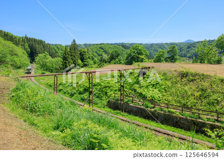 長年支撐著田地的紅褐色水管，一列飯山線柴油火車從水管下方駛過。 125646594