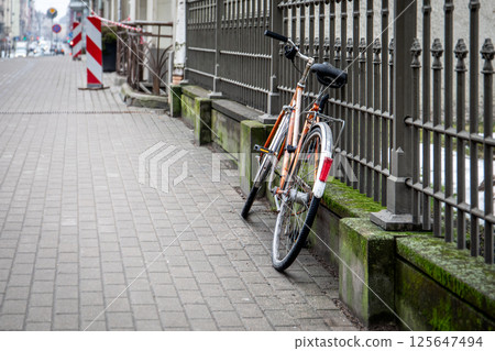 Abandoned Bicycle Leaning Against Fence on Paved Sidewalk Abandoned Bicycle Leaning Against Fence on Paved Sidewalk 125647494