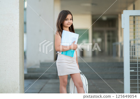 Little child with brown hair in skirt standing in street on her way to school carrying backpack 125649054