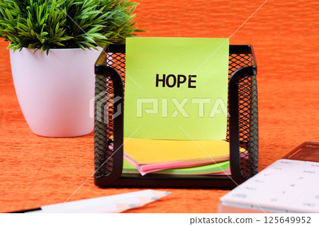 A black mesh organizer holds bright sticky notes, with a prominent note reading hope. A small green plant adds a touch of nature on a vibrant orange background 125649952