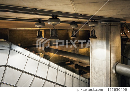Industrial looking architecture, metal lamps shine over tubes, concrete walls and pillars in modern train station. Blurred people going down the escalator stairs Industrial looking architecture, metal lamps shine over tubes, concrete walls and pillars in modern train station. Blurred people going down the escalator stairs 125650496