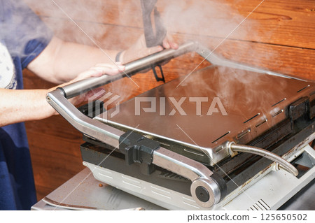 Detail on woman hands holding closed electric grill handle, smoke above as food is grilled 125650502