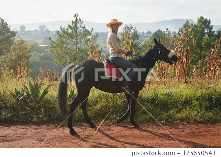 Young woman in shirt and straw hat, riding black horse in the park on sunny morning, blurred background with corn plants, houses and trees Young woman in shirt and straw hat, riding black horse in the park on sunny morning, blurred background with corn plants, houses and trees 125650541