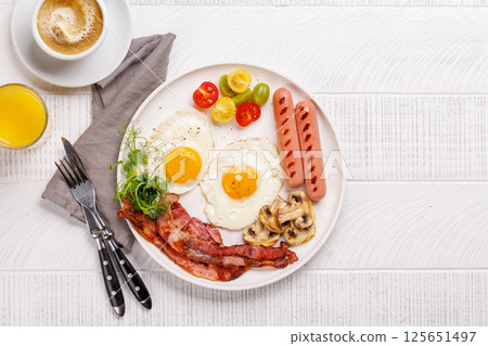 A classic breakfast plate featuring sunny-side-up eggs, crispy bacon, sausages, and fresh tomatoes A classic breakfast plate featuring sunny-side-up eggs, crispy bacon, sausages, and fresh tomatoes 125651497