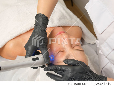 Close-up portrait of a beautiful aged woman during microneedling with dermapen from a beautician. Close-up portrait of a beautiful aged woman during microneedling with dermapen from a beautician. 125651810