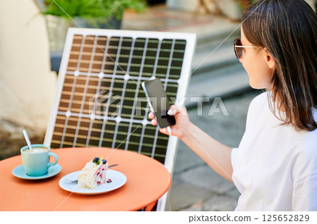Young woman charging her smartphone from solar panel while relaxing. Pretty girl drinking coffee and checking her phone next to solar battery. Conscious female choosing sustainable lifestyle. 125652829