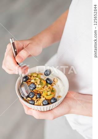 Bowl of granola with greek yogurt, kiwi and blueberries and spoon in hands of woman 125652844
