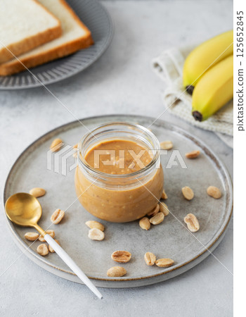 Peanut butter in glass jar on light  background with salted nuts, banana and bread toasts. 125652845
