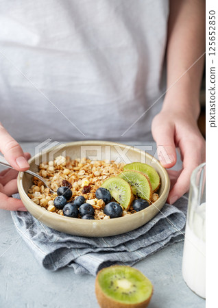 Hand holding bowl of homemade baked granola with kiwi and blueberries on a light background Hand holding bowl of homemade baked granola with kiwi and blueberries on a light background 125652850