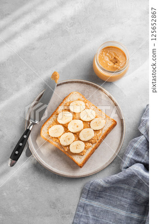 Peanut butter toast with banana and knife on a plate on a light background with nuts 125652867