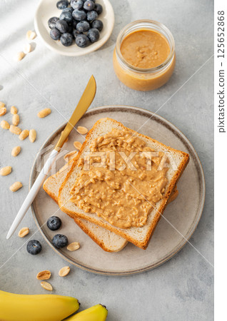 Peanut butter toast on a plate on a light background with banana, blueberry and morning shadow. 125652868