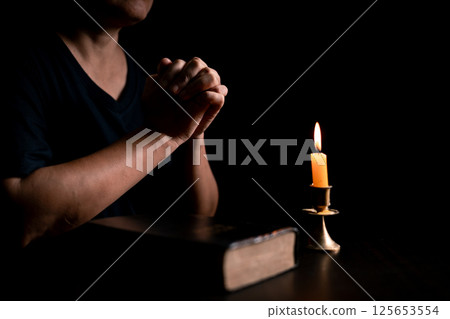In the dark, hand clasped in prayer, illuminated by the soft candlelight, as black flames of candle fire flickered gently. candle, pray, hand, prayer, black, fire, flames, candlelight, light, dark. 125653554