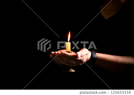 In the dark, hand clasped in prayer, illuminated by the soft candlelight, as black flames of candle fire flickered gently. candle, pray, hand, prayer, black, fire, flames, candlelight, light, dark. 125653559