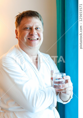 A man starts his morning with a glass of clean drinking water standing near the window 125653571